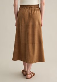 Jupe maxi en cuir marron avec une taille élastique, présentant une texture lisse et des détails de coutures, associée à des sandales à lanières marron.