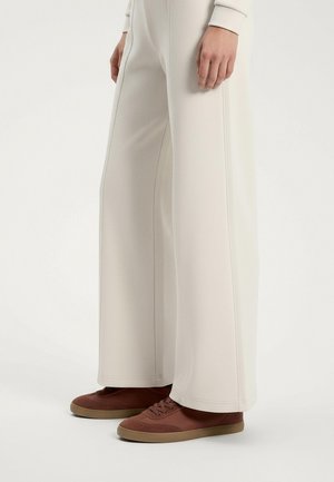 Široke, kremaste hlače od teksturiranog materijala, uparene s smeškim patikama koje imaju baršunaste i glatke detalje.