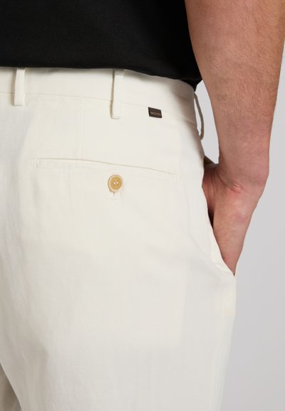 Nahaufnahme einer Person, die weiße Hosen mit einer geknöpften Gesäßtasche und einem schwarzen Oberteil trägt, die Hand in der vorderen Tasche steckend.