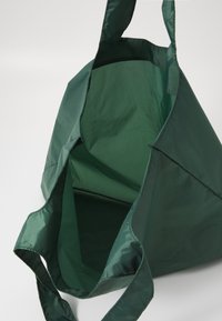 Borsa a tracolla verde realizzata in tessuto leggero e lucido. Caratterizzata da un ampio interno con una fodera a motivo a griglia e due manici robusti.