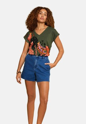 Frau mit lockigem Haar, die ein grünes V-Ausschnitt-Hemd mit orangefarbenem Tiermuster und blaue Jeansshorts trägt, steht mit einer Hand in der Tasche.