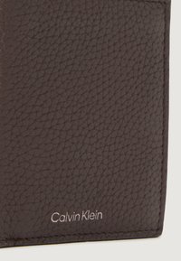 Cartera de cuero marrón texturizado con el logo de "Calvin Klein" en plata. Diseño delgado con costuras limpias y acentos minimalistas.