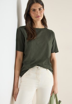 Jeune femme aux longs cheveux bruns portant un t-shirt vert foncé et un pantalon blanc, debout contre un mur clair, tenant un tissu vert.