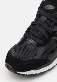 Chaussure de sport noire avec une tige en mesh et des accents en daim. Lacets épais et semelle durable avec des détails blancs sur la semelle intermédiaire.