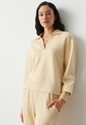 Vrouw met lang donker haar, gekleed in een crèmekleurige trui met kraag en bijpassende broek, staand met één hand in de zak tegen een witte muur.