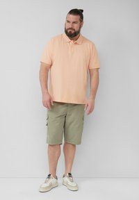 Korte mouwen perzikkleur polo, olijfgroene cargoshorts met zakken, en witte sneakers met beige accenten. Neutrale achtergrond.