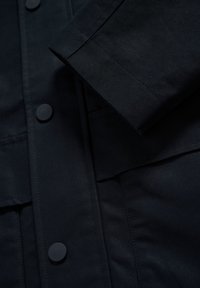 Temno modra jakna iz gladkega blaga, z velikimi gumbi, žepom na krpo in klasičnim ovratnikom z ravnim robom ter prilagojenimi rokavi.