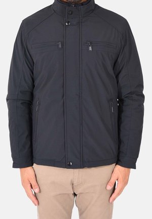 Man draagt een zwarte jas met ritssluiting, twee borstzakken en twee zijzakken met ritssluiting, gecombineerd met beige broek, staat met ontspannen handen.