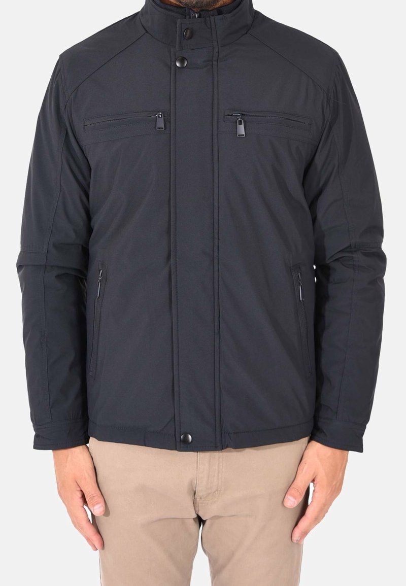 Homme portant une veste noire zippée avec deux poches sur la poitrine et deux poches latérales zippées, associée à un pantalon beige, debout les mains détendues.