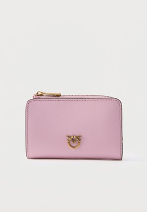 Portefeuille en cuir rose clair avec fermeture éclair dorée et emblème décoratif en or représentant deux oiseaux tenant un anneau à l'avant.