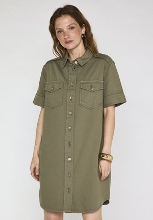 Femme portant une robe chemise à manches courtes vert olive avec deux poches poitrine, des boucles d'oreilles anneaux et des bracelets en or au poignet droit.