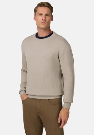Beige texturový svetr s kulatým výstřihem a žebrovanými manžetami. Nosený přes tmavě modrou košili, v kombinaci s hnědými kalhotami.