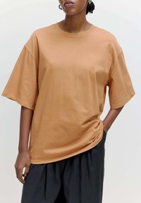 Losse, kortaf T-shirt in een lichtbruine stof met een gladde textuur. Ronde halslijn en een ontspannen silhoutte. Gekleed met donkere broeken.