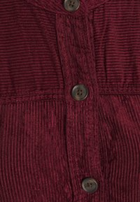 Gros plan sur un tissu en velours côtelé rouge profond avec des rainures verticales et trois boutons gris foncé à quatre trous cousus sur un vêtement.