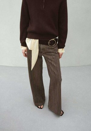 Persona que lleva pantalones anchos de rayas marrón, suéter marrón oscuro, puños de camisa beige, cinturón negro con hebilla dorada y zapatos negros abiertos en la punta.