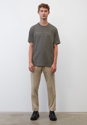 Olivgrünes T-Shirt mit Frontdruck, kombiniert mit beigen Slim-Fit-Hosen und schwarzen Schnürschuhen. Einfaches Design, weiche Stoffstruktur.