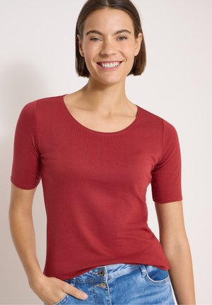Femme souriante aux cheveux bruns courts, portant un t-shirt rouge à manches courtes et un jean bleu, la main gauche dans la poche.