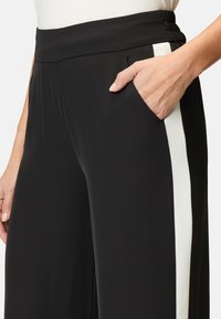 Schwarze Hosen mit breitem Bündchen, seitlichem Taschen und einem weißen seitlichen Streifen. Der Stoff wirkt glatt und leicht elastisch.
