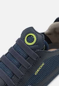 Zapatilla azul marino con parte superior de malla, acentos de plástico y ojales verdes. Presenta correas elásticas y una suela texturizada para agarre. Marca Geox presente.