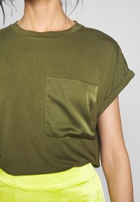 Olivově zelené bavlněné tričko s texturovanou saténovou kapsou, krátkými ohrnutými rukávy a volným střihem; spárováno s jasně žlutou sukní.