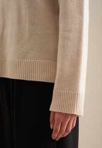 Beige gebreide trui met detaillering zoals een geribbelde boord en mouwen, een zachte textuur en een losse pasvorm, waarbij een gladde mouw gedeeltelijk zichtbaar is.
