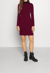Robe bordeaux à manches longues avec un motif texturé, coupe ajustée et col montant, associée à des bottines noires à lacets ornées de clous.