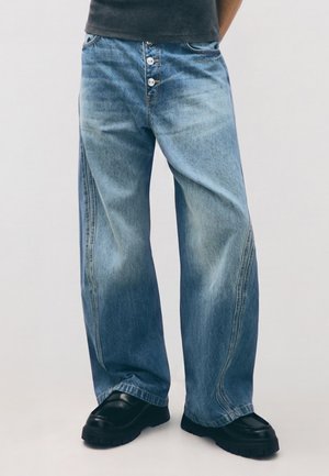 Person trägt weite, ausgebleichte blaue Jeans mit Knopfleiste und schwarze klobige Schuhe, steht vor einem schlichten hellen Hintergrund.