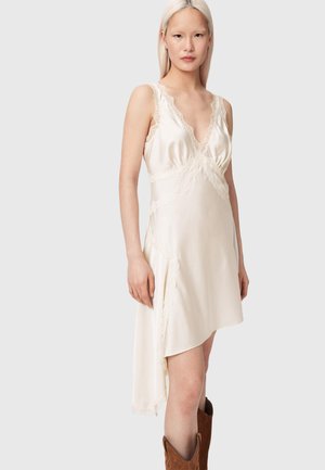 Kobieta w kremowej, satynowej, asymetrycznej sukience z koronkowym obszyciem i brązowych kowbojskich butach, stojąca na tle jednolitego szarego tła.