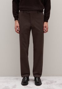 Pantaloni marroni su misura in tessuto strutturato, con chiusura a bottone e tasche laterali. Abbinati a scarpe nere, in piedi su una superficie chiara.