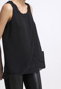 Top senza maniche nero con scollo rotondo, tessuto fluido e orlo asimmetrico, abbinato a pantaloni neri slim. Texture morbida, design leggero.