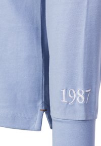 Light blue long-sleeve shirt made of cotton, featuring a side split, embroidered "1987" in white on the sleeve, and minimal stitching details.