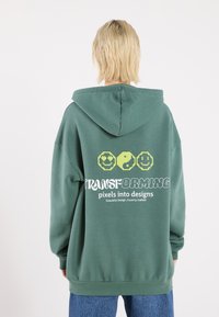 Felpa verde con cappuccio; il retro presenta faccine sorridenti gialle in stile pixelato e testo. Tessuto morbido, vestibilità rilassata, polsini e orlo a coste.