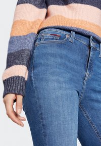 Pruhovaný pletený svetr v pastelových barvách, zkombinovaný s modrými džíny. Džíny mají zip, kapsy a standardní pas.