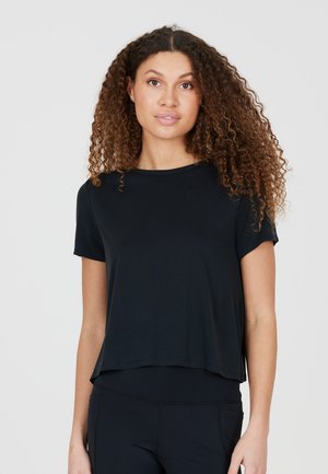 Mujer con cabello rizado y castaño que lleva una camiseta negra holgada de manga corta y pantalones negros sobre un fondo liso y claro.