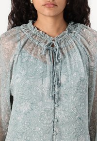 Hellbluse mit floralem Muster aus transparenter Stoff, ausgestattet mit einem gerafften Ausschnitt, einer Bindeschließe und dekorativen Knöpfen entlang der Vorderseite.