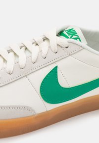 Weiße Sportschuhe aus Leder mit grünen Akzenten. Sie verfügen über einen auffälligen grünen Nike-Swoosh, eine strukturierten grauen Überlagerung und eine Gummisohle.