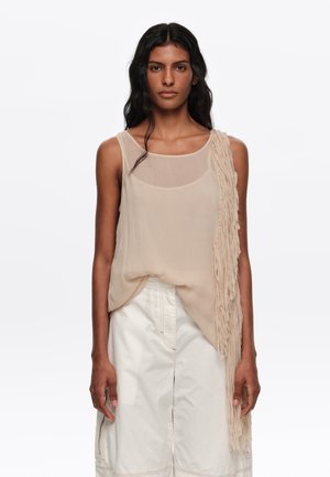 Mujer con pelo largo, oscuro y ondulado, viste una blusa sin mangas beige con detalle de flecos en un lado y pantalones blancos de talle alto, de pie frente a un fondo blanco.
