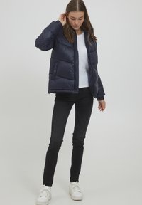 Giubbotto imbottito blu navy con chiusura a zip, polsini a coste e tasche laterali, indossato sopra una maglietta bianca e abbinato a jeans skinny neri e sneakers bianche.