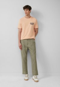 Perzikkleurig T-shirt met zwarte bedrukte tekst op de linkerborst; olijfgroene broek; witte sneakers met beige accenten; eenvoudig ontwerp, casual pasvorm.