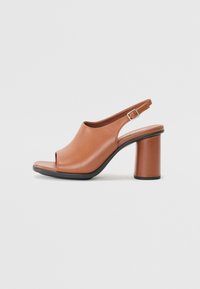 Zapato abierto en la parte delantera, de cuero marrón, con talón bloque, textura suave y detalle de hebilla ajustable. La suela contrasta con la parte superior.