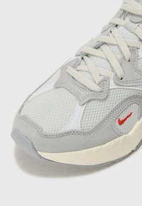 Grijze sportieve schoen van mesh en suede materialen, met gestructureerde oppervlakken, een crèmekleurige zool en een klein rood logoaccent aan de zijkant.