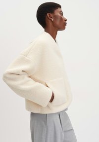 Giacca di pile color crema con una vestibilità ampia, spalle scese e una tasca frontale. Presenta una texture morbida e un design minimalista.