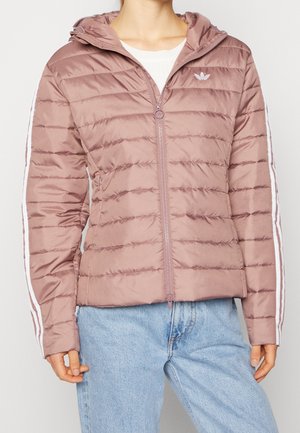 Femme portant une veste matelassée mauve avec capuche et bandes blanches sur les manches, associée à un jean en denim bleu clair.