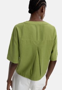 Zielona bluzka z krótkim rękawem o luźnym kroju, z plisą z tyłu i okrągłym dekoltem. Wykonana z lekkiego materiału.