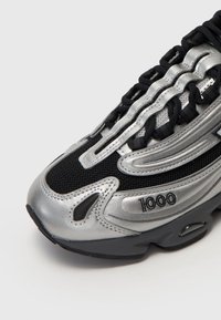 Srebrno-czarny sportowy but z siateczkową cholewką, charakterystycznym falistym wzorem i wyściełanym kołnierzem. Posiada logo "1000" oraz fakturowane akcenty.