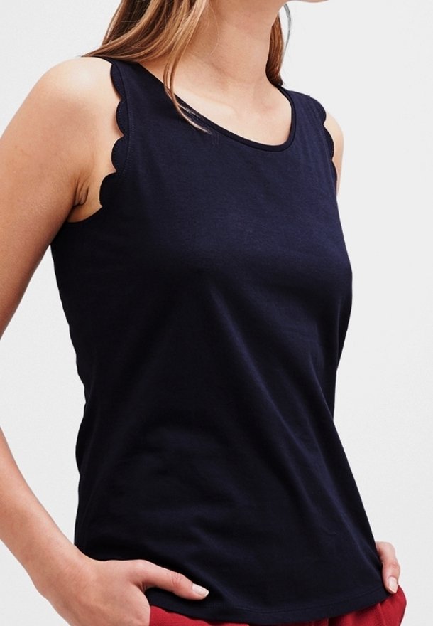Femme portant un haut noir sans manches avec des bords festonnés aux épaules, associé à un pantalon rouge, debout les mains dans les poches.