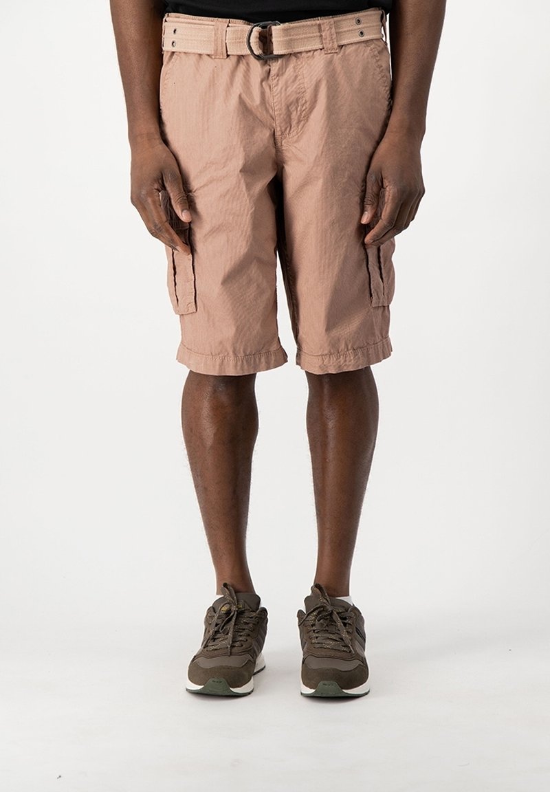 Homme portant un short cargo marron clair avec une ceinture en tissu et des baskets marron foncé, debout devant un fond blanc uni.
