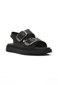 Sandalias de cuero negro con doble hebilla metálica, diseño de punta abierta, suelas resistentes y detalles texturizados en la parte superior.