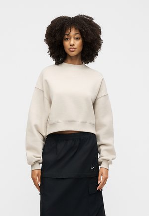 Femme aux cheveux bouclés portant un sweat-shirt court beige et une jupe noire avec un logo Nike, debout devant un fond clair uni.