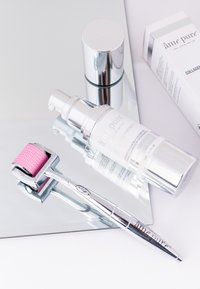 Rosa dermaroller med teksturert hode, sølvhåndtak og reflekterende overflater, sammen med to hvite og sølv hudpleieflasker og en eske.